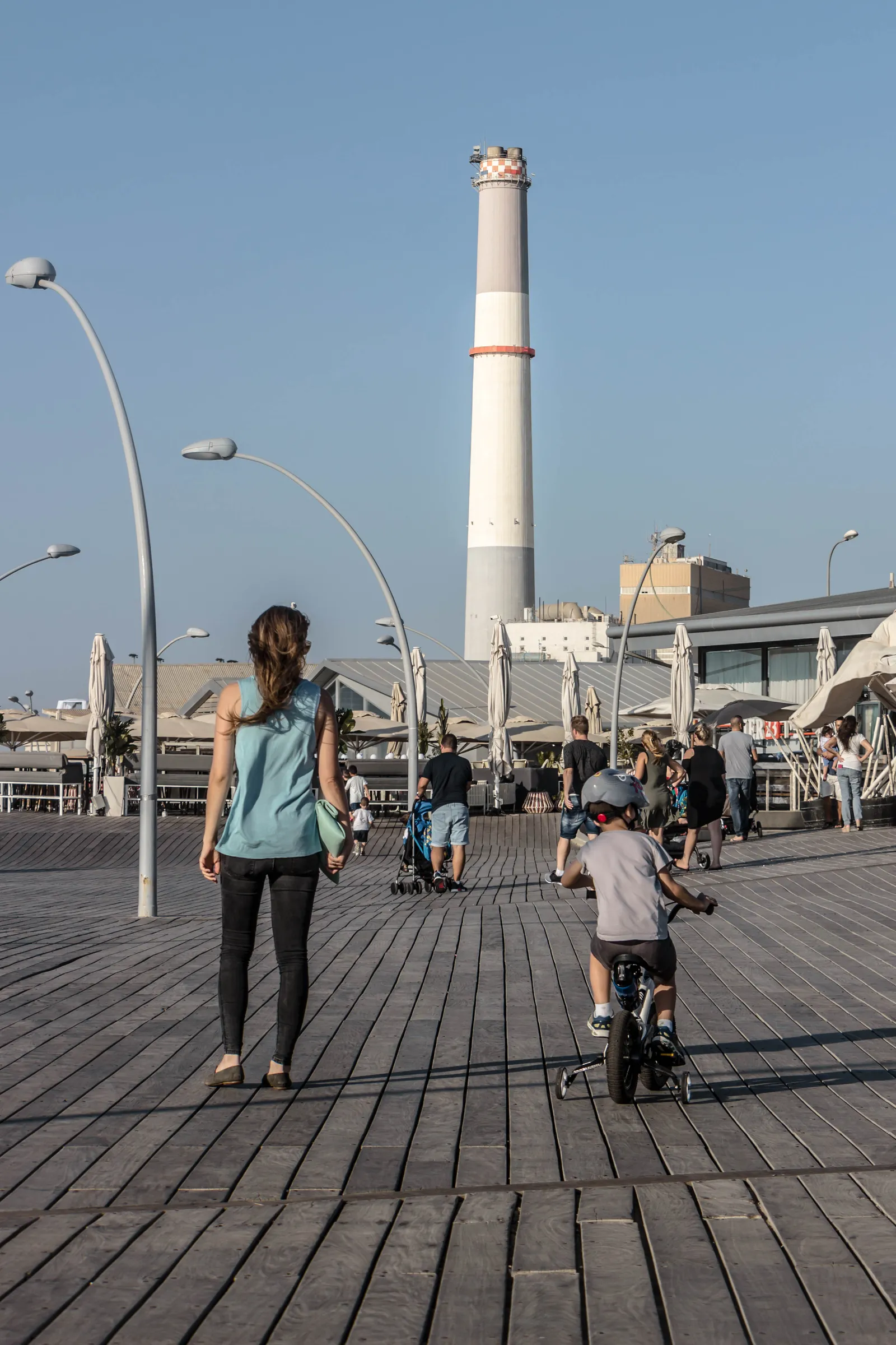 The timber boardwalk at the old Tel Aviv port, its planks laid in long curves, with a woman walking and a child on a balance bike ahead of her and the banded chimney of the power station rising beyond the hangar roofs
