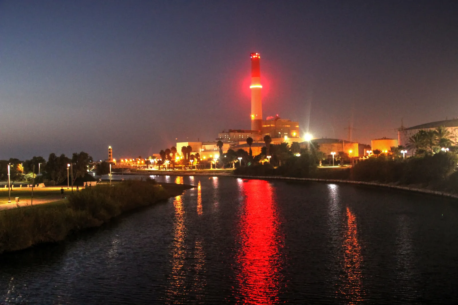 The Reading power station at the mouth of the Yarkon in Tel Aviv after dark, its tall chimney lit red above the low-lit plant buildings and reflected down the length of the river