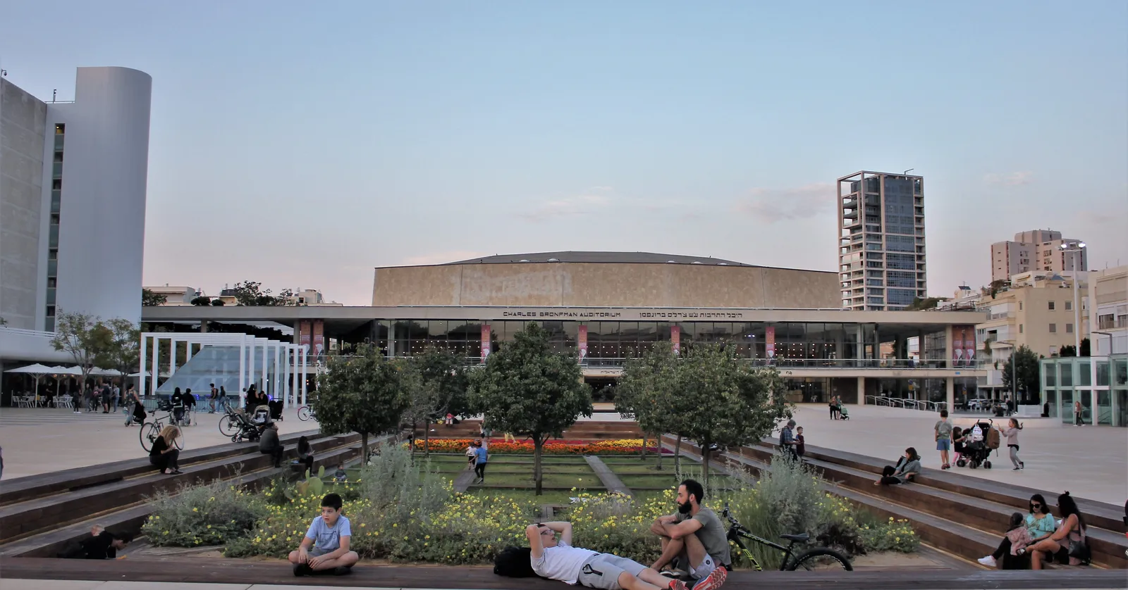 The Charles Bronfman Auditorium on Habima Square in Tel Aviv, a long low concert hall with a ribbed pale facade, seen across the square's sunken garden with people sitting on the stepped stone edges in front of it