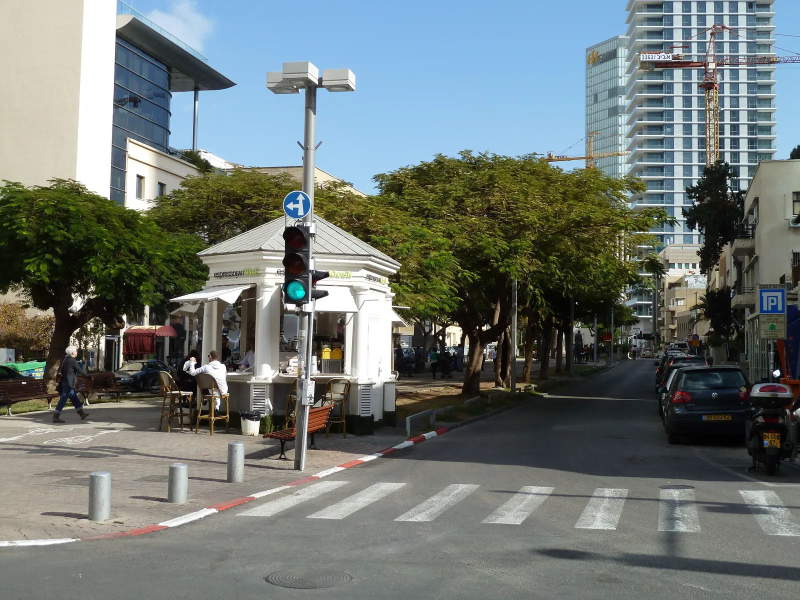The small white hexagonal kiosk at the corner of Rothschild Boulevard and Herzl Street in Tel Aviv, with a scalloped roof, a serving window and a few pavement tables under the trees beside it