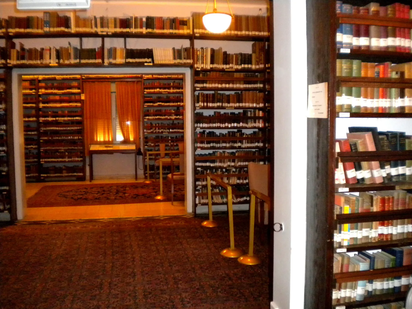 Inside Ben-Gurion House in Tel Aviv, floor-to-ceiling shelves of books lining every wall of two connected rooms, a patterned rug on the floor and a lit reading table at the far end