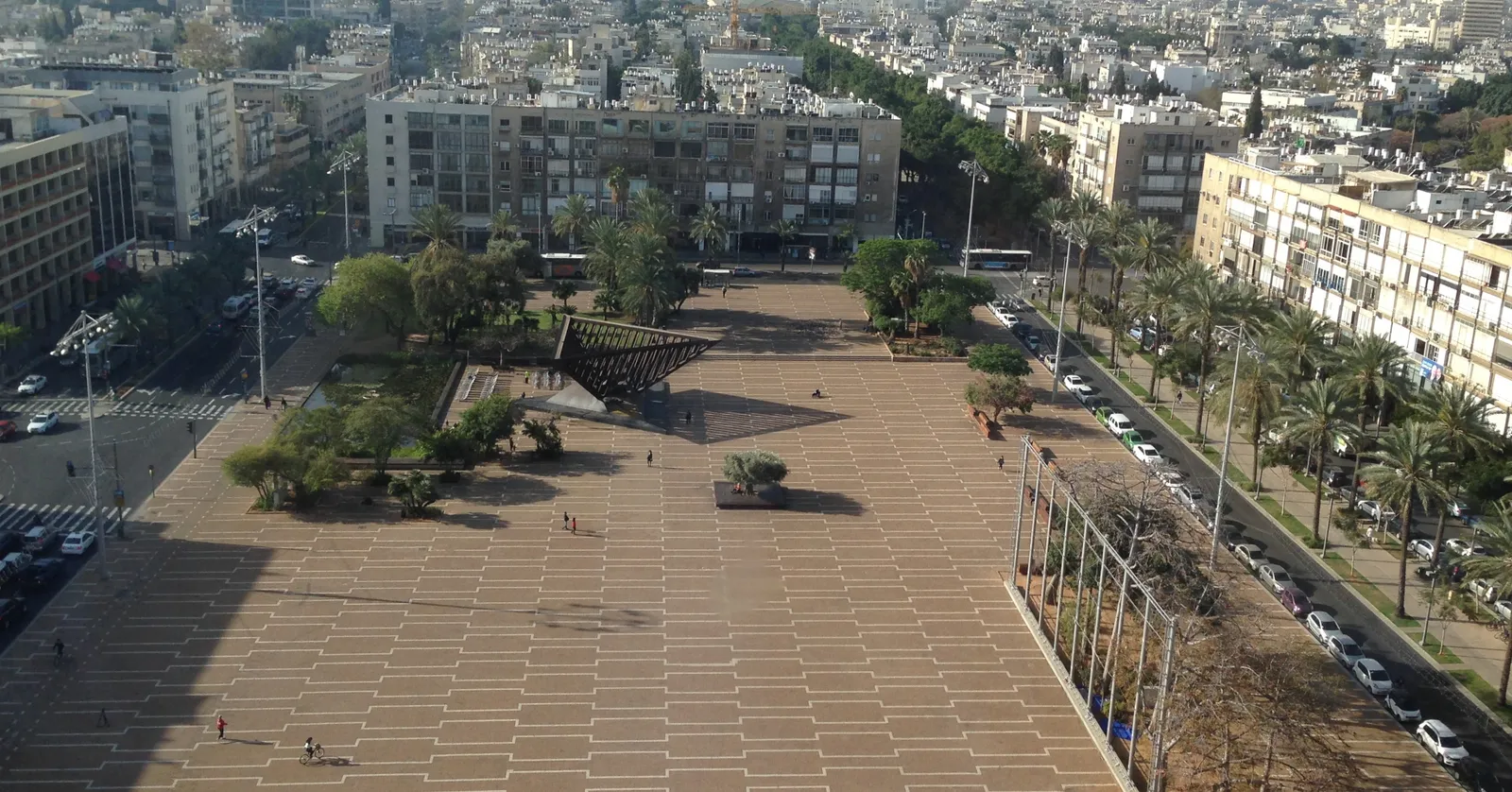 Rabin Square in Tel Aviv seen from above, a vast expanse of pale paving stones ringed by low apartment blocks and palm trees, with the towers of the city rising in the haze behind