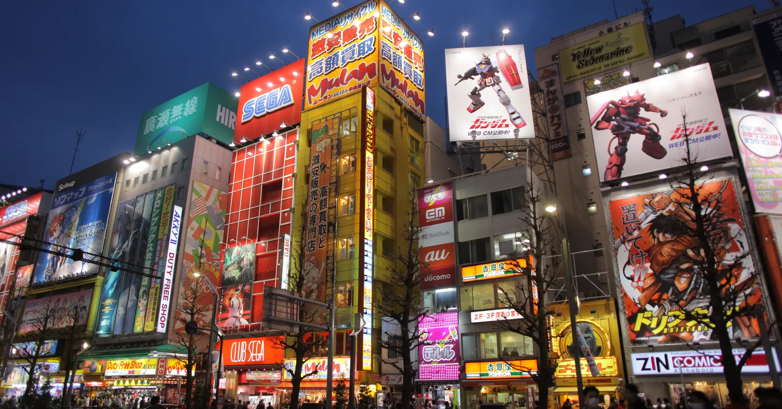 Chuo-dori in Akihabara, Tokyo, at dusk, a row of tall narrow shop buildings covered top to bottom in lit signs and giant anime and game billboards, with shoppers on the pavement below