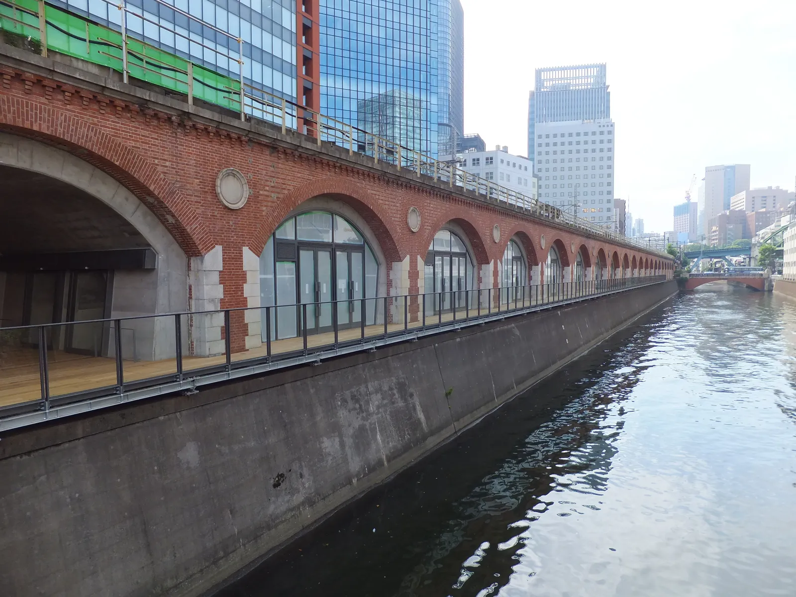 The red-brick arches of the old Manseibashi railway viaduct in Tokyo running along the Kanda River, glass shopfronts set into each arch and a riverside deck along the water, with office towers behind