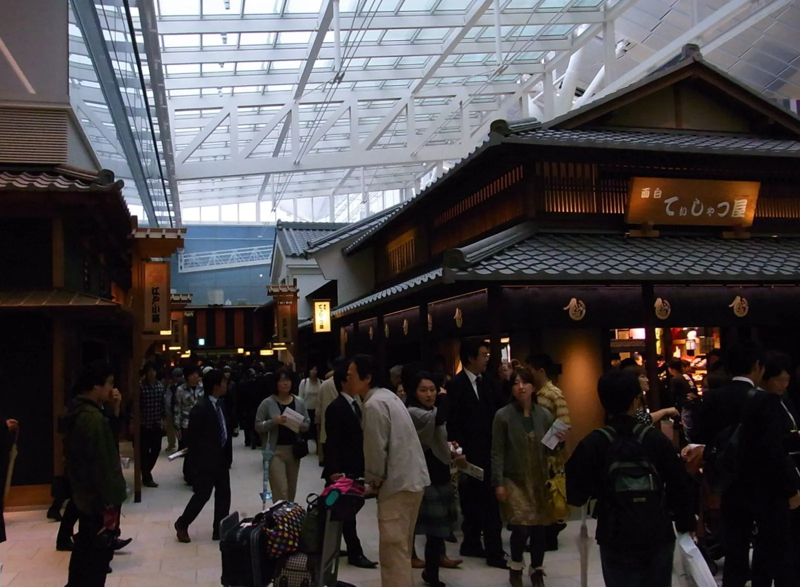 The Edo-style shopping street inside Terminal 3 at Haneda Airport, timber-fronted shops with tiled roofs and hanging signs under the terminal's glass roof, with travellers and their luggage walking past