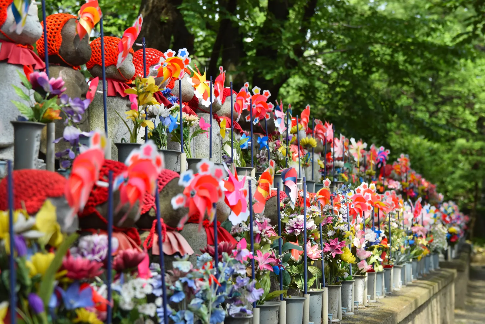 A long row of small stone Jizo statues at Zojo-ji, each wearing a red knitted cap and bib, with colourful plastic pinwheels and flowers set in front of them under the trees