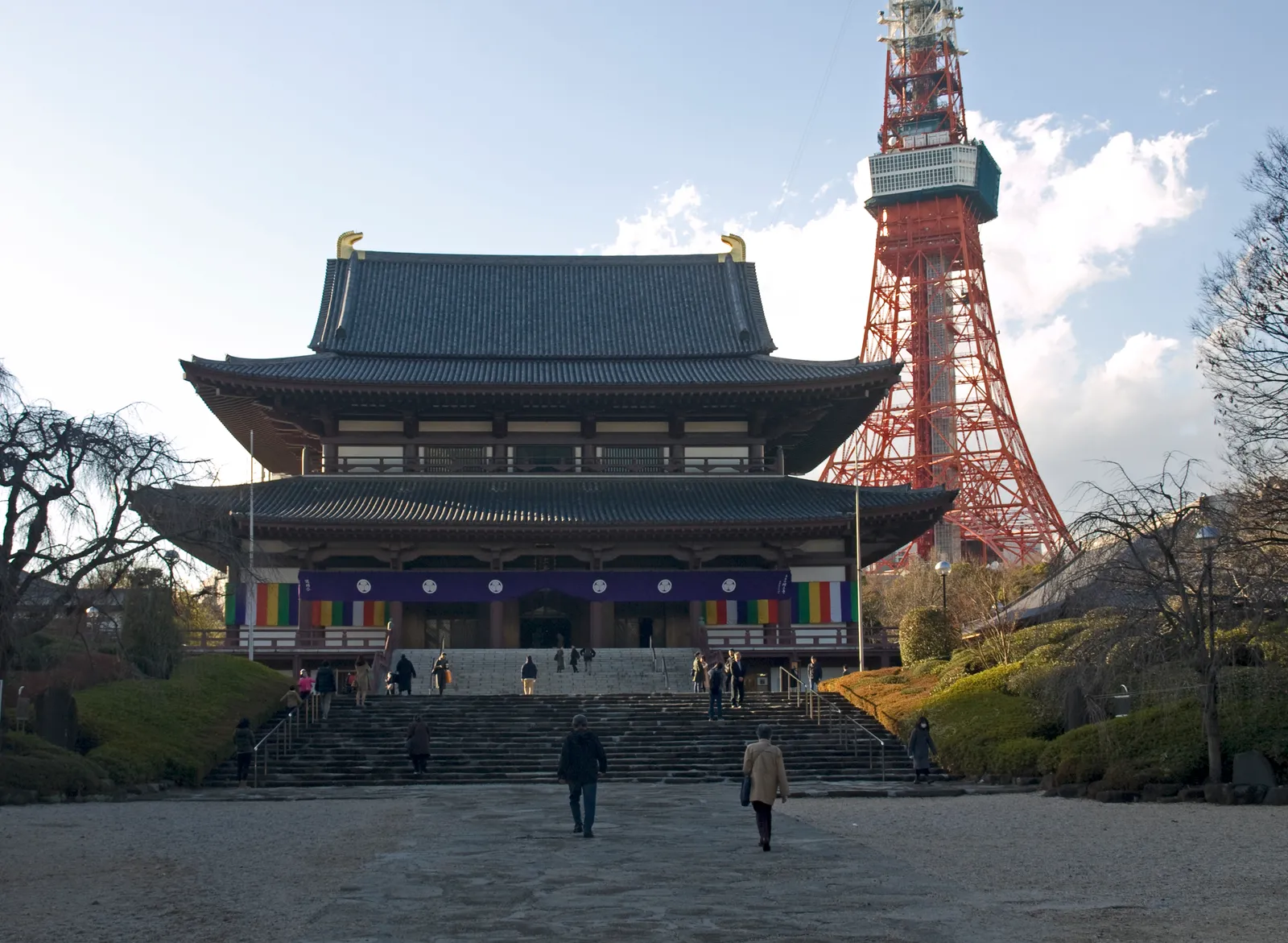The main hall of Zojo-ji in Tokyo, a two-tiered temple building with a dark tiled roof at the top of a broad flight of stone steps, with the red and white lattice of Tokyo Tower rising directly behind it