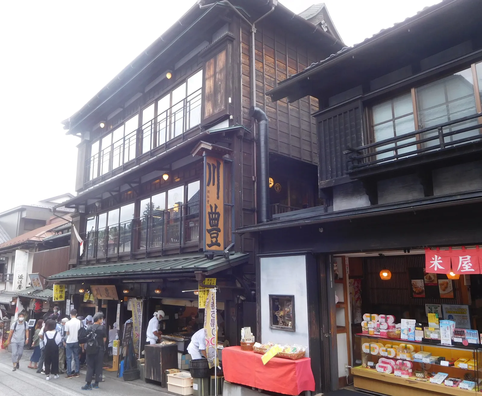 Kawatoyo Honten on Narita's Omotesando, a three-storey dark timber eel restaurant with a hanging wooden signboard and an open shopfront at street level, people queuing and browsing along the pavement outside