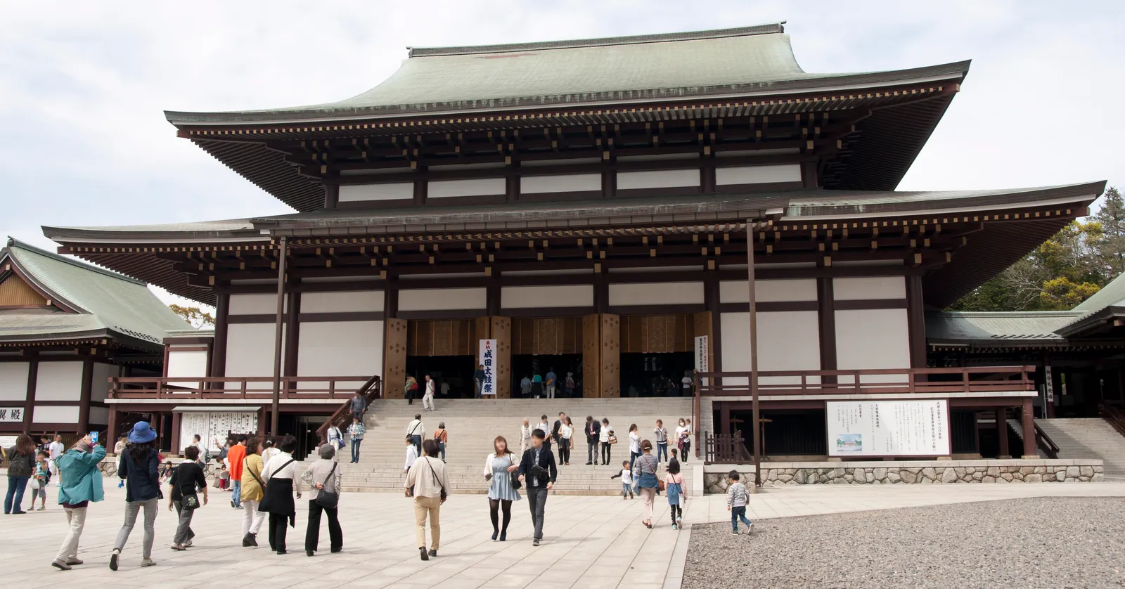 The Great Main Hall at Naritasan Shinshoji in Narita, a broad two-tiered wooden hall under a green copper roof with a wide flight of stone steps up to its open doors and visitors crossing the paved forecourt in front