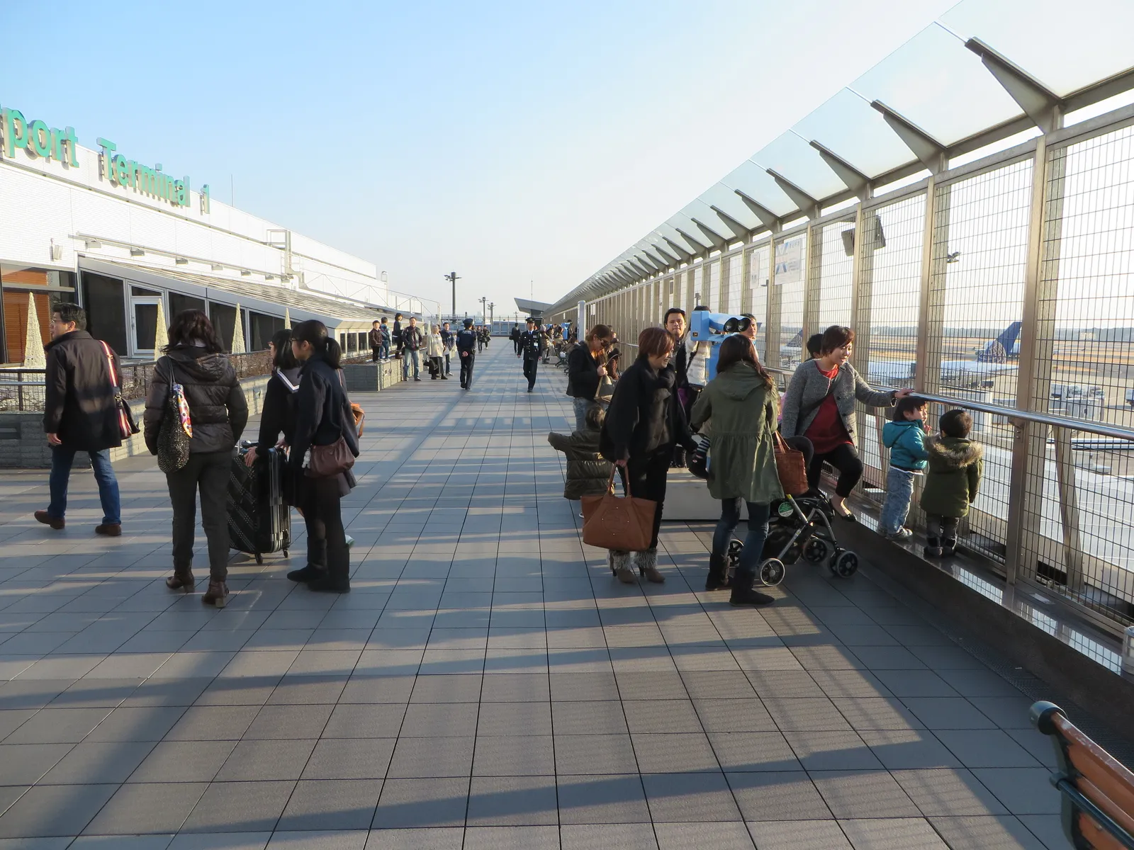 The open-air observation deck on top of Terminal 1 at Narita International Airport, a long paved terrace behind a tall glass and steel railing with families looking out at parked aircraft in the late afternoon sun