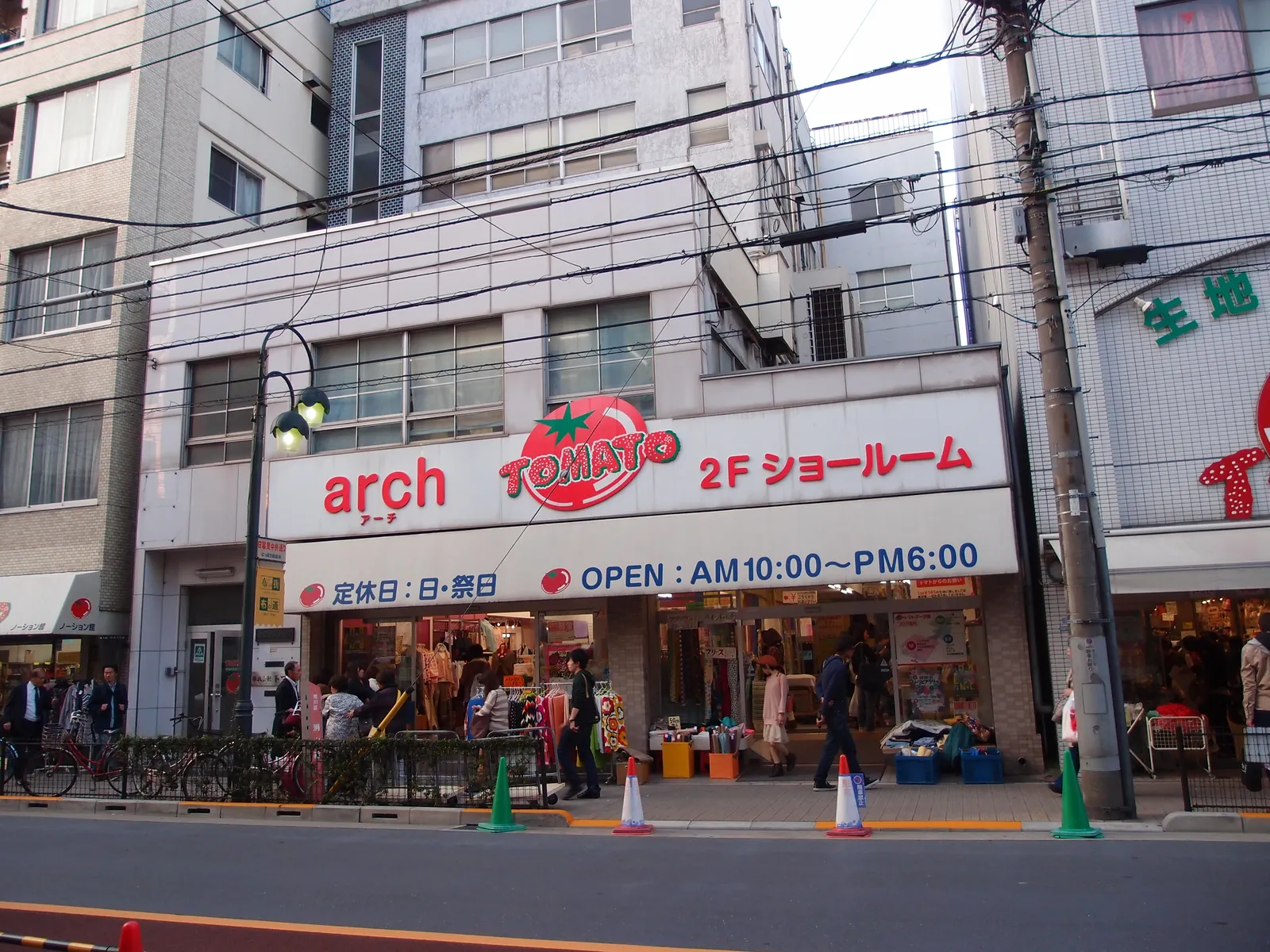 The Tomato fabric store on Nippori's textile street in Tokyo, a white multi-storey shopfront with a red tomato sign, bolts and rolls of cloth stacked on the pavement outside and shoppers browsing them