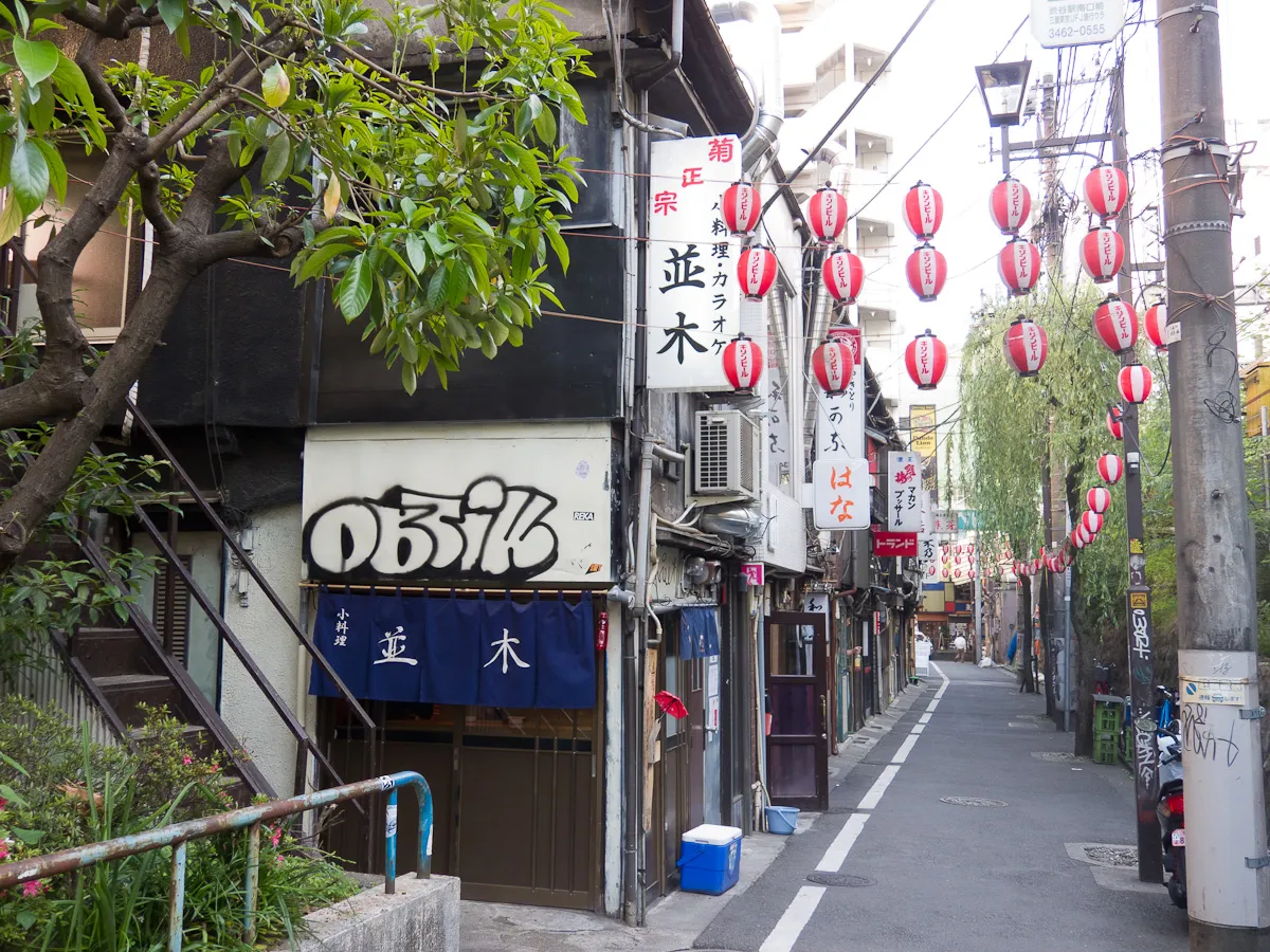 Nonbei Yokocho in Shibuya, a narrow lane of tiny two-storey bars with wooden fronts, hand-lettered signs and strings of red paper lanterns running down both sides beside the railway