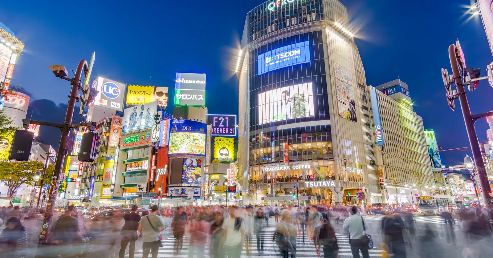 The Shibuya scramble crossing in Tokyo at dusk, crowds blurred in motion across the zebra stripes in every direction under a deep blue sky, with lit video screens and signs covering the buildings around the junction