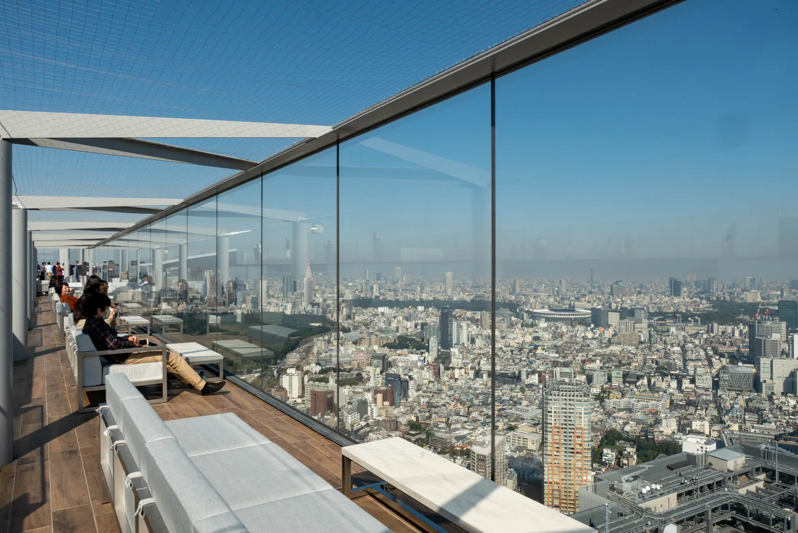 The open-air roof of Shibuya Sky in Tokyo, a timber deck with low white couches running along a tall glass parapet, and the city spreading flat to the horizon beyond it on a clear day