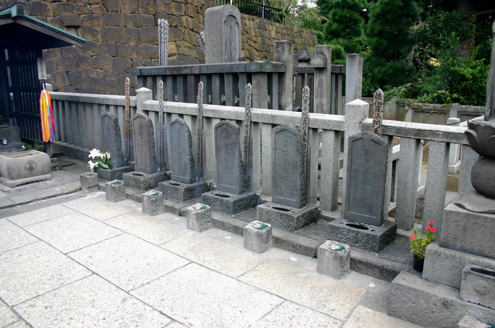 The graves of the 47 ronin at Sengaku-ji in Tokyo, rows of narrow upright stone markers inside low stone and wooden fences, with incense holders at their feet and flowers left on several of them