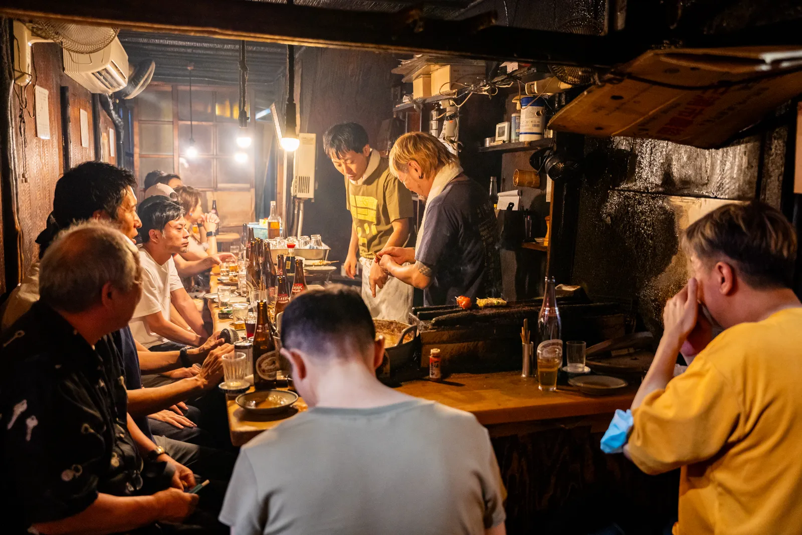 Customers packed shoulder to shoulder along the counter of a tiny yakitori bar in Omoide Yokocho in Shinjuku, cooks working at the grill behind it under bare bulbs and smoke-darkened walls