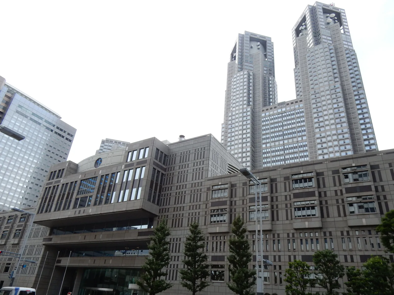 The twin towers of the Tokyo Metropolitan Government Building in Shinjuku, two grey latticed towers rising from a broad lower block, seen from street level behind a row of trees