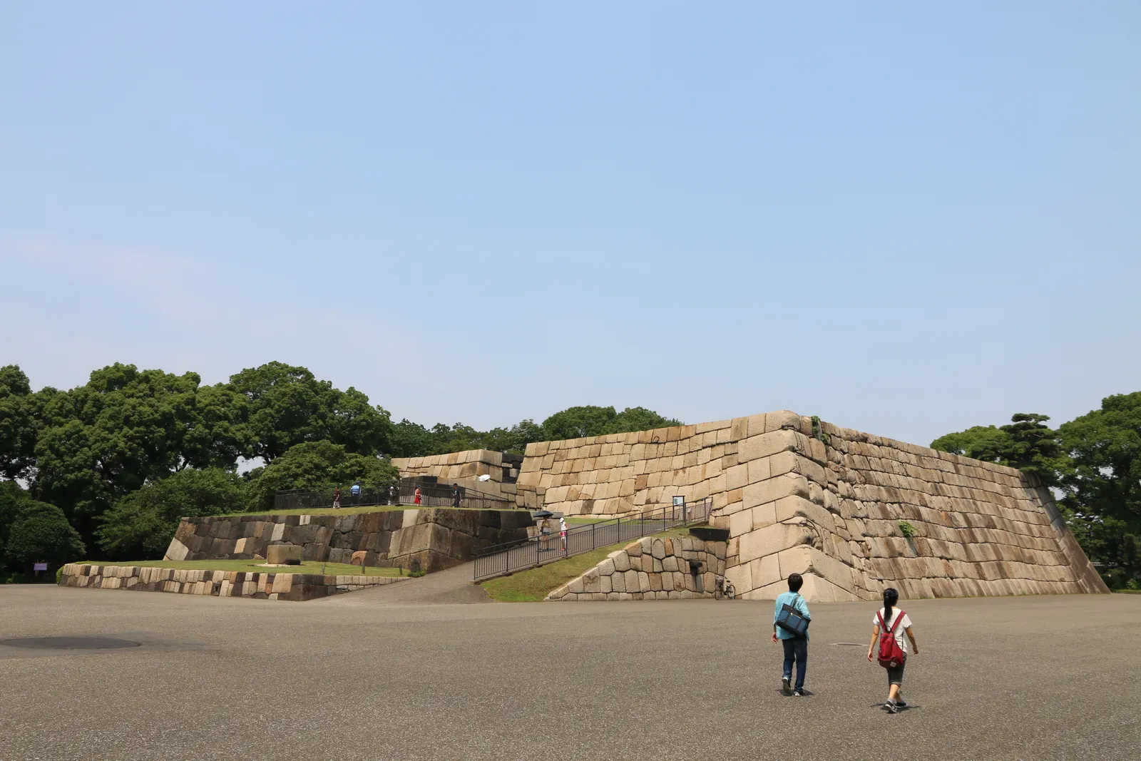 The stone base of Edo Castle's keep in the Imperial Palace East Gardens, a broad flat-topped platform of huge fitted granite blocks with a ramp up one side, two visitors crossing the open gravel ground in front of it