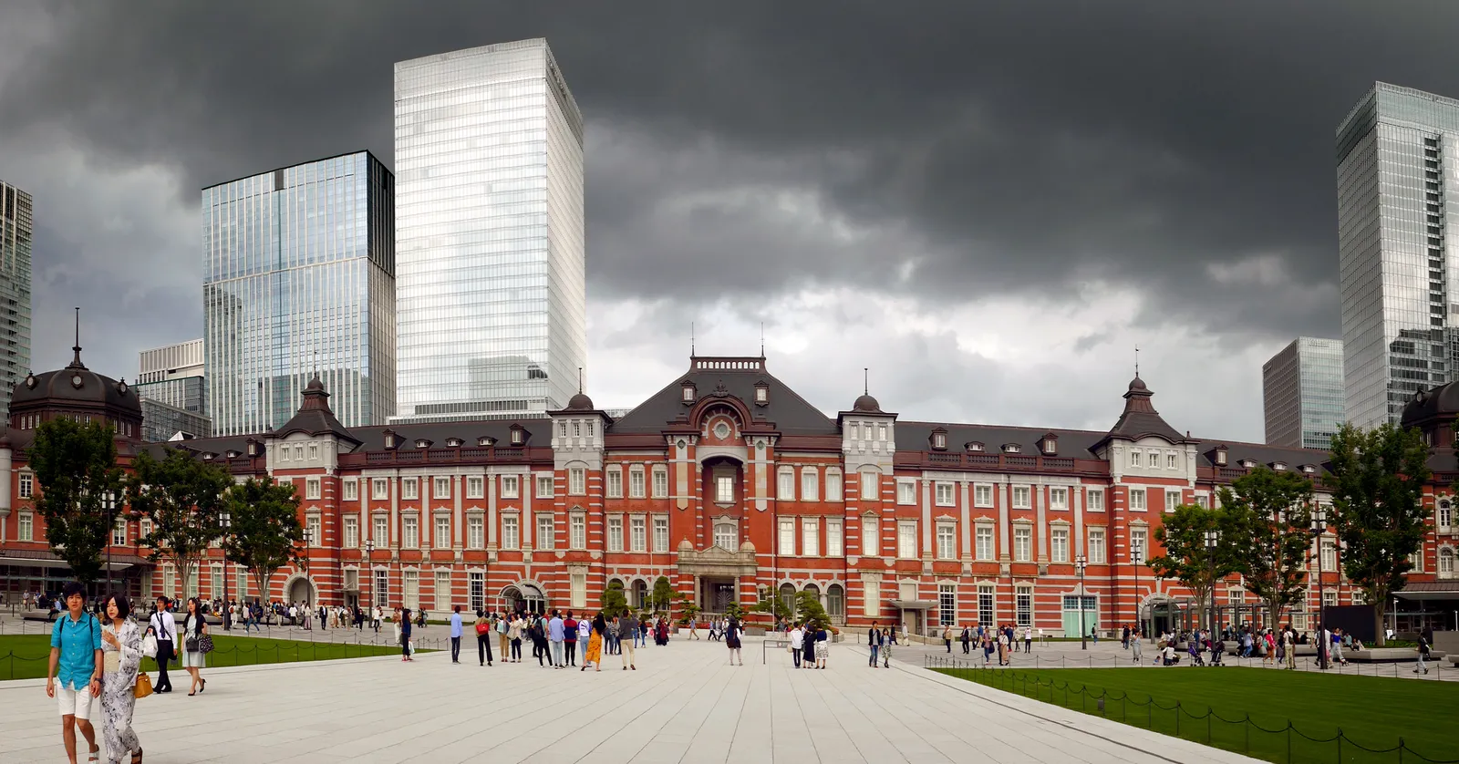 The red-brick Marunouchi front of Tokyo Station, a long three-storey building with white stone banding and a dark domed roof at each end, seen across the wide paved plaza in front of it with glass office towers rising behind