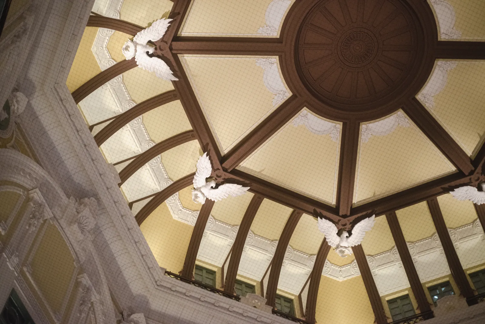 Looking straight up into the north dome of Tokyo Station, a cream and ochre octagonal ceiling divided by dark ribs, with white plaster eagles spreading their wings at the base of each rib