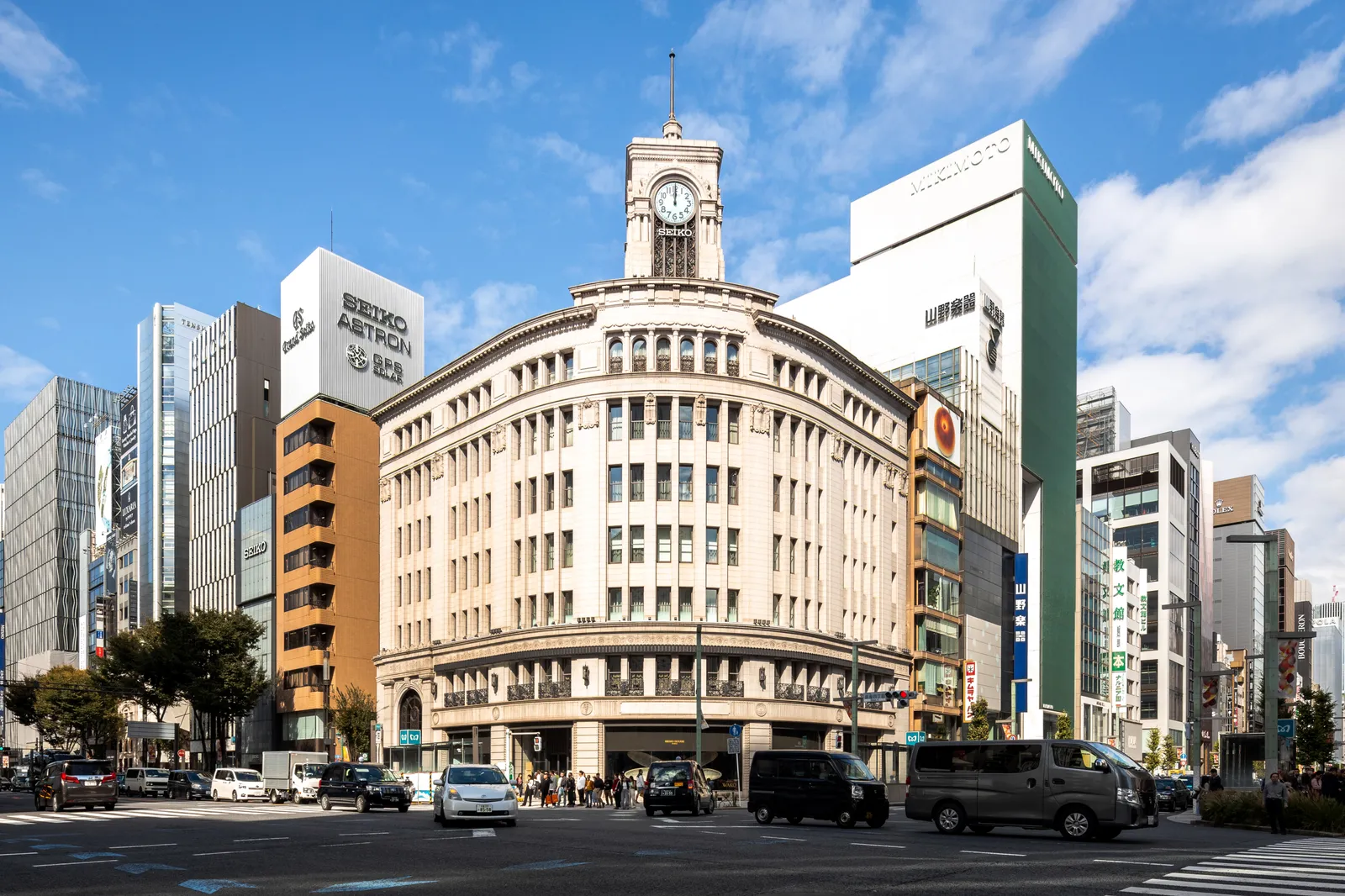 The Wako building on the corner of Ginza's main crossing in Tokyo, a curved cream stone facade rising to a clock tower, with shopfronts and glass towers lining the wide street on either side under a blue sky
