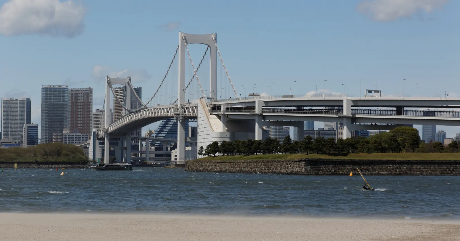 The sand beach of Odaiba Seaside Park in Tokyo in front of calm bay water, with the white towers and cables of the Rainbow Bridge spanning across to the city's office towers on the far shore