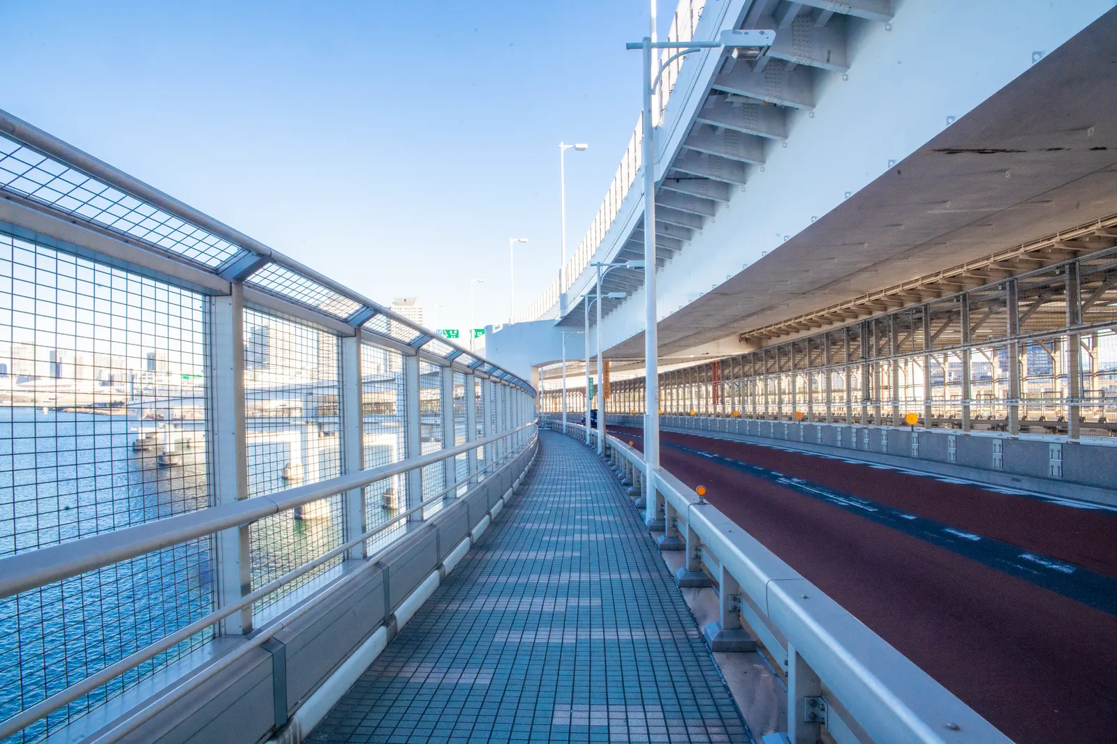 The pedestrian walkway along the side of Tokyo's Rainbow Bridge, a long straight path behind a tall mesh railing with the open water of the bay below and the bridge deck structure overhead