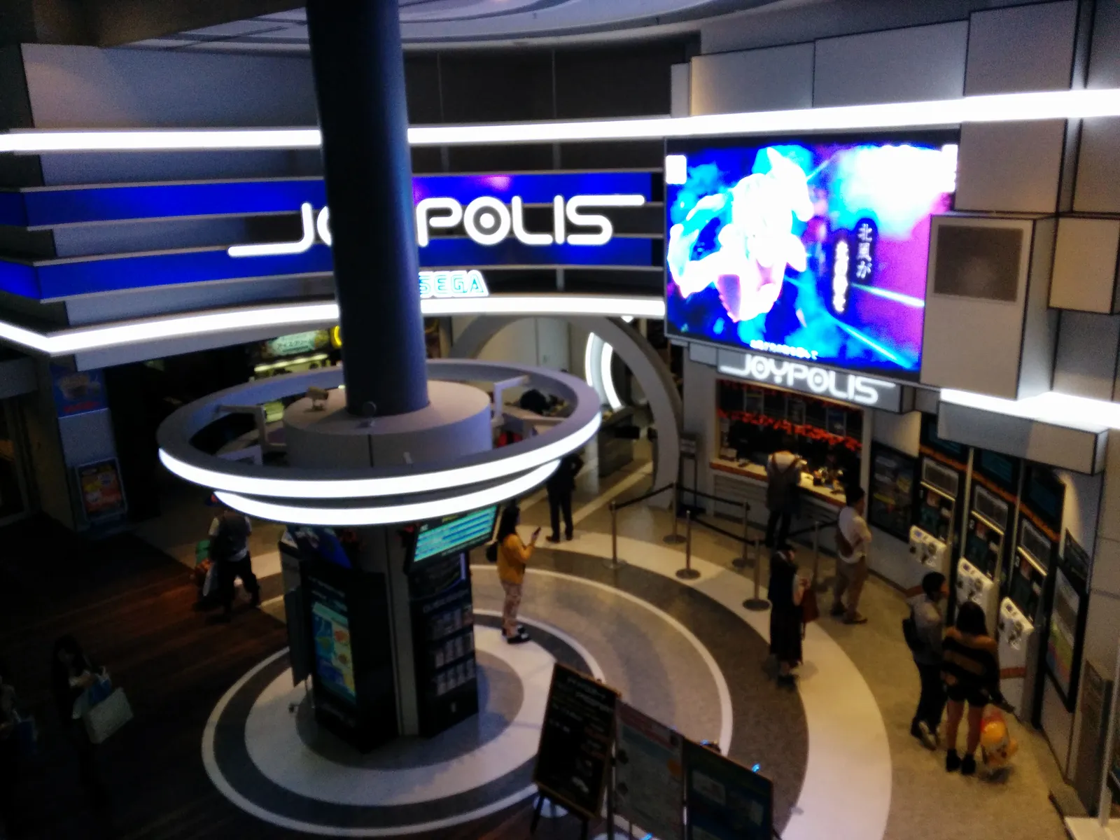 The entrance hall of Tokyo Joypolis on Odaiba, a dark circular atrium with ring-shaped neon lights, a large video screen and the illuminated Joypolis logo above the ticket gates