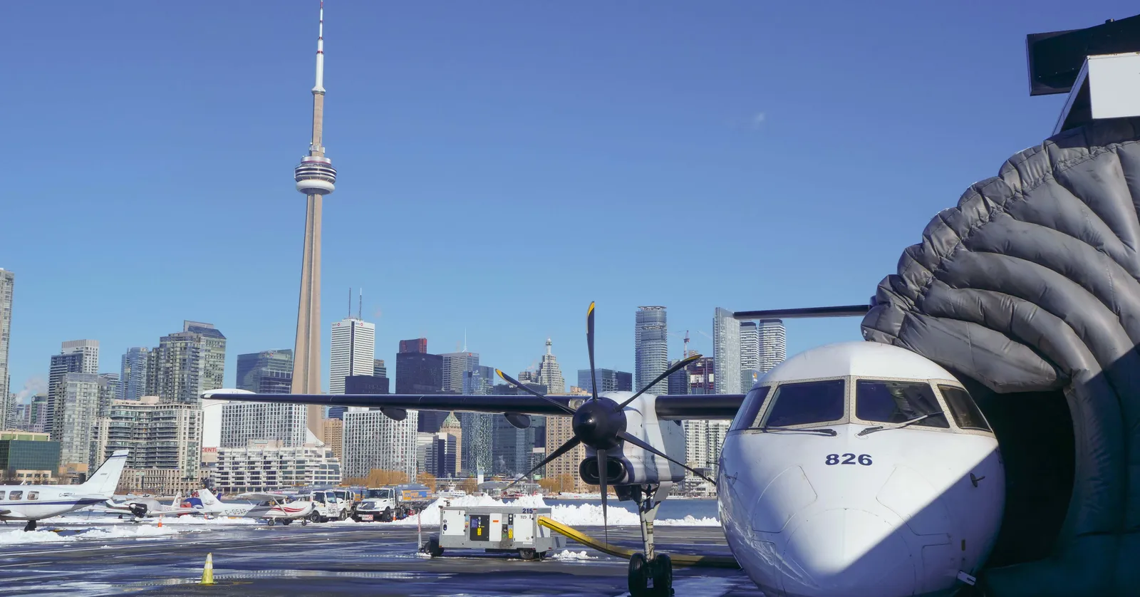 A white turboprop airliner parked at a jet bridge at Billy Bishop Toronto City Airport, with the downtown Toronto skyline and the CN Tower rising directly behind it across the water