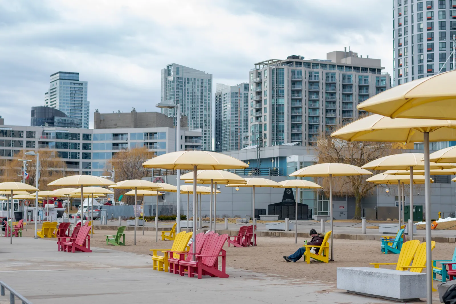 Rows of large yellow umbrellas over sand at HTO Park in Toronto, with brightly painted wooden deck chairs underneath them and residential towers along the street behind