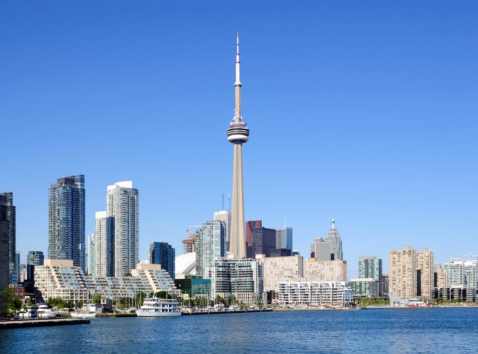 Toronto's inner harbour seen from the water, with the terraced waterfront apartment blocks of Queens Quay along the shore, moored boats in front of them and the CN Tower behind