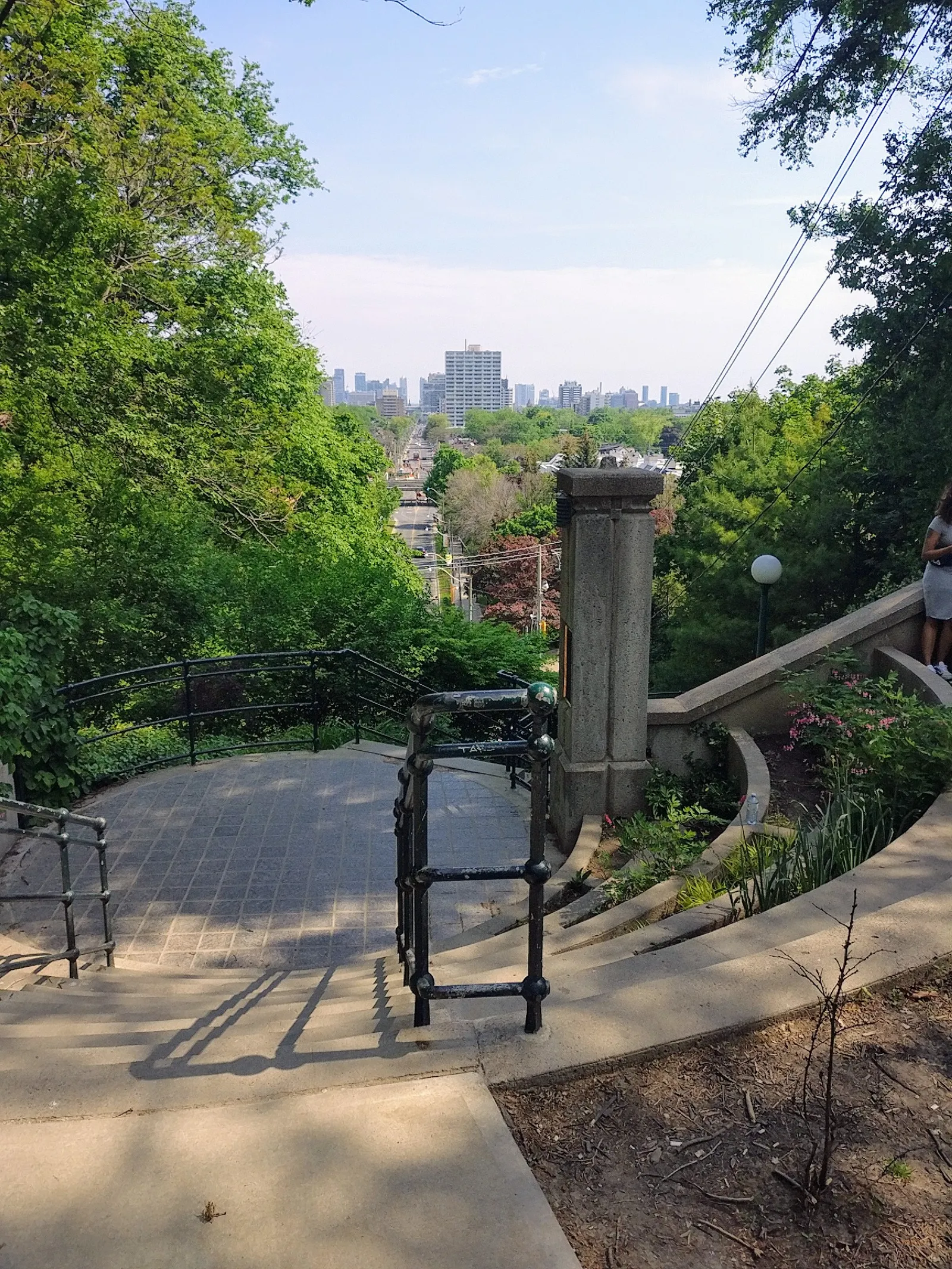 The top of the Baldwin Steps in Toronto, a public stone staircase descending an escarpment between iron handrails and trees, with the downtown skyline visible in the distance