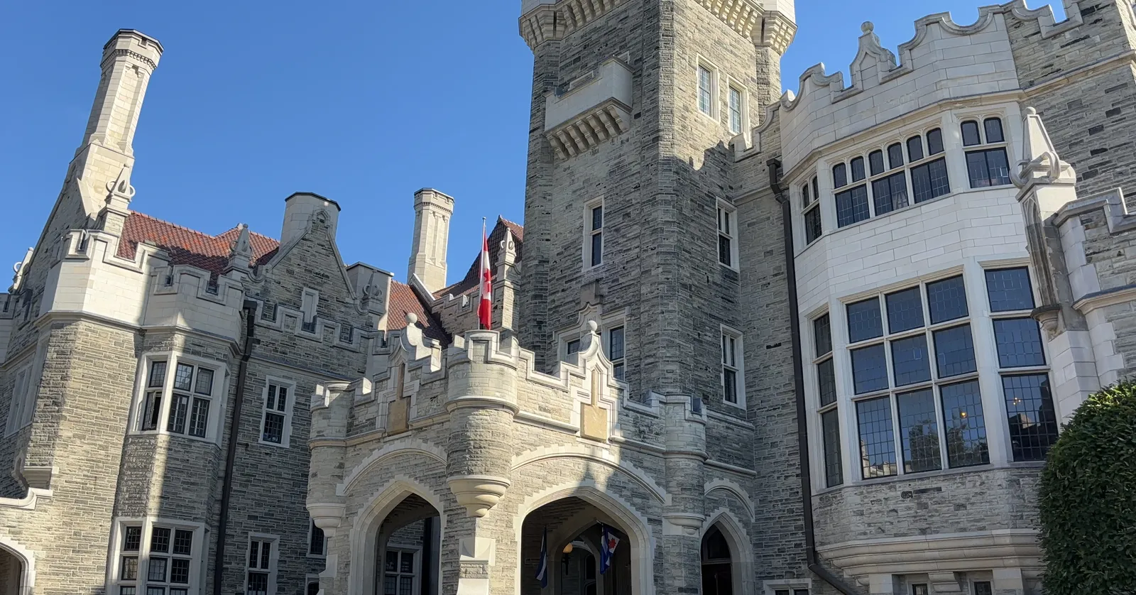 The grey stone front of Casa Loma in Toronto seen from below, with castellated towers, mullioned bay windows and flags flying above the arched entrance