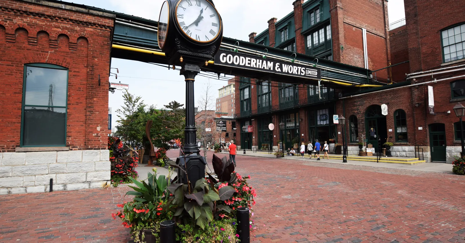 A brick-paved lane in Toronto's Distillery District lined with red-brick warehouses, a black and gold Gooderham and Worts sign spanning the street overhead and planters of red flowers in the foreground