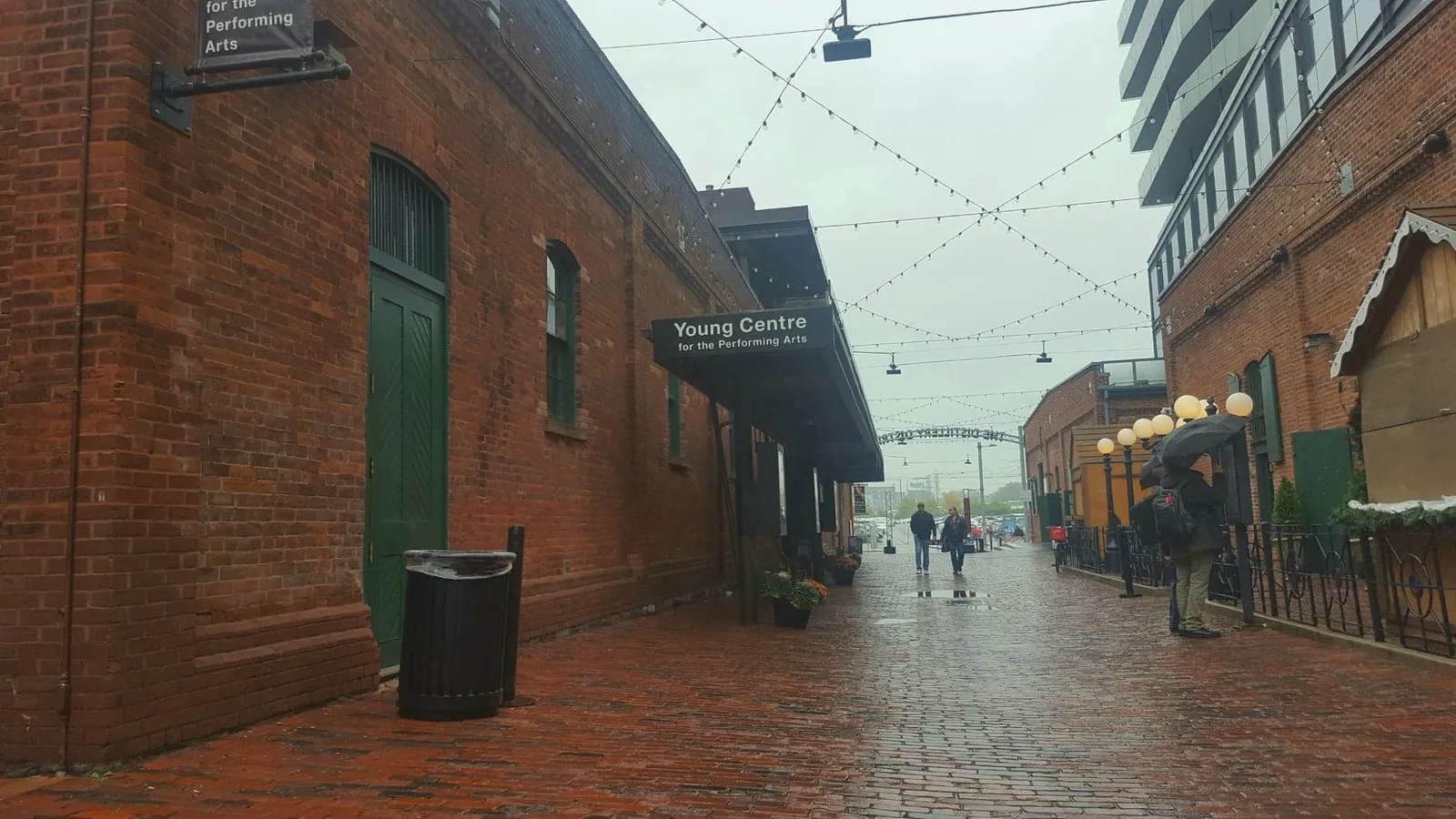 A wet brick lane in Toronto's Distillery District with a Young Centre for the Performing Arts sign projecting from a red-brick wall, string lights overhead and people walking in the distance