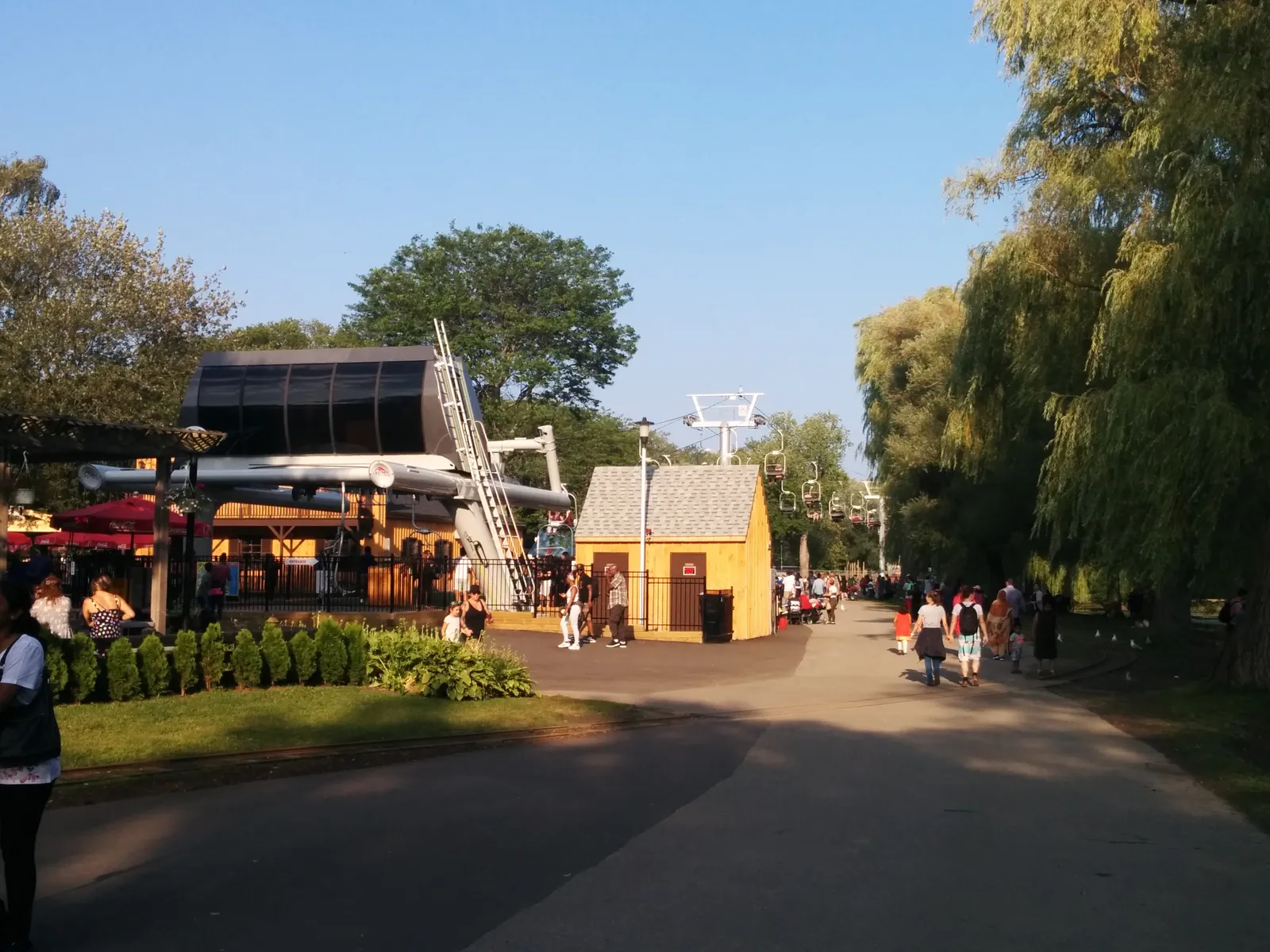 A path through Centreville amusement park on Centre Island in Toronto, with a yellow ride building, a windmill structure, willow trees and families walking in the sun