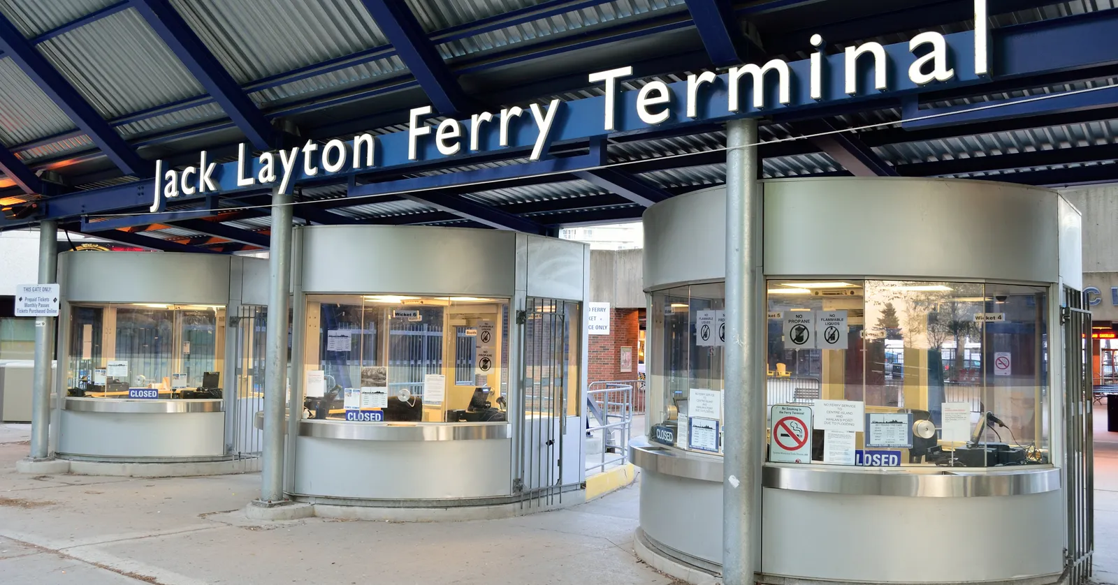 The ticket booths under the canopy at the Jack Layton Ferry Terminal in Toronto, curved glass kiosks lit from inside with the terminal's name spelled out on the fascia above them