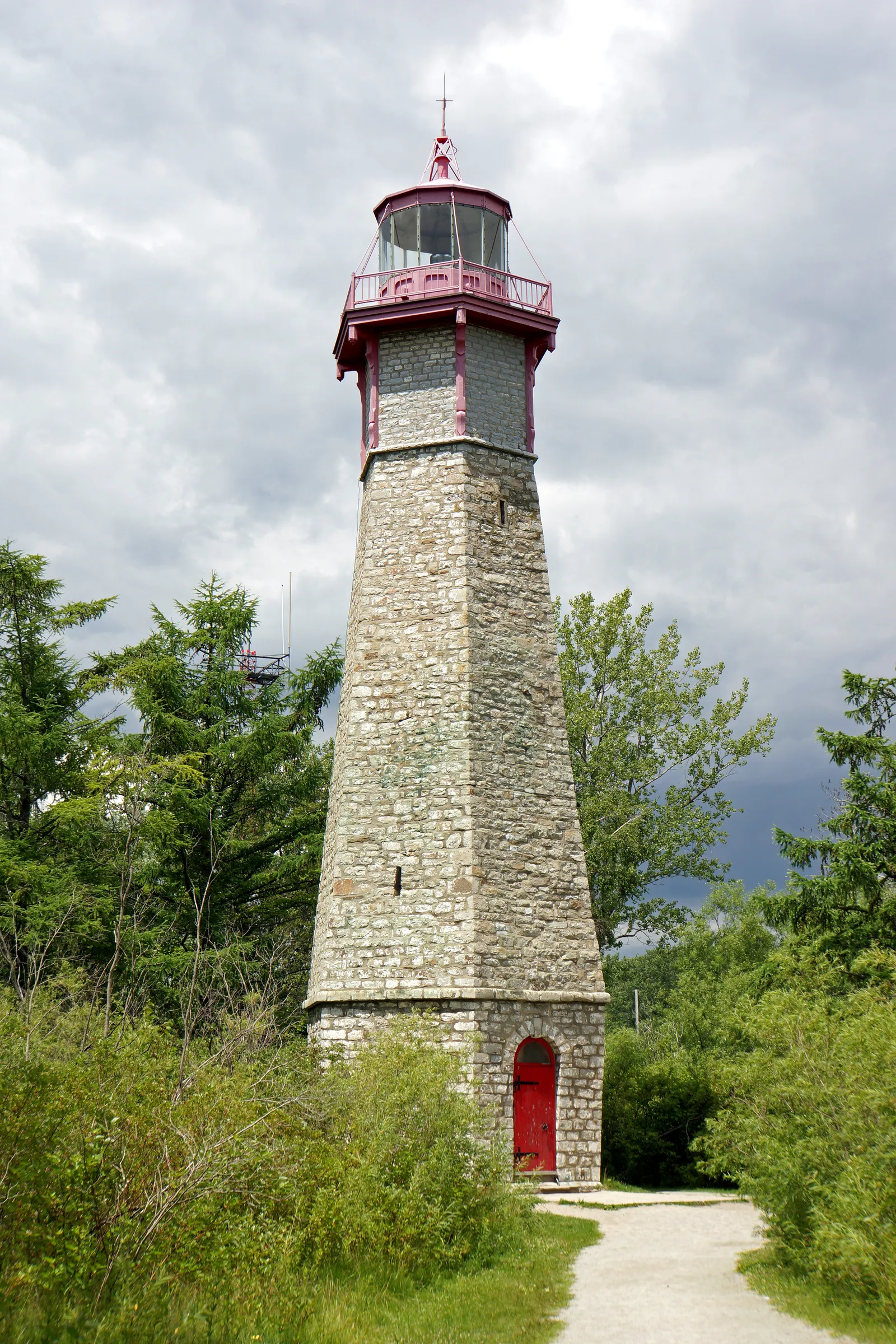 The Gibraltar Point Lighthouse on the Toronto Islands, a tapering stone tower with a red-painted lantern and gallery at the top, standing among trees under a grey sky