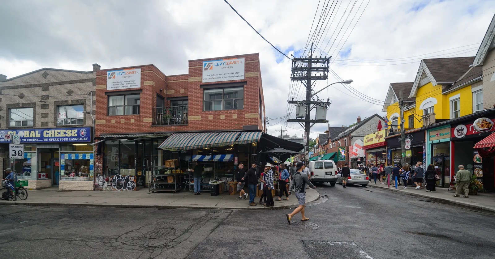 A street corner in Kensington Market, Toronto, with a cheese shop and a green-awninged produce stall in a converted brick house, painted Victorian shopfronts opposite and people crossing the road between them