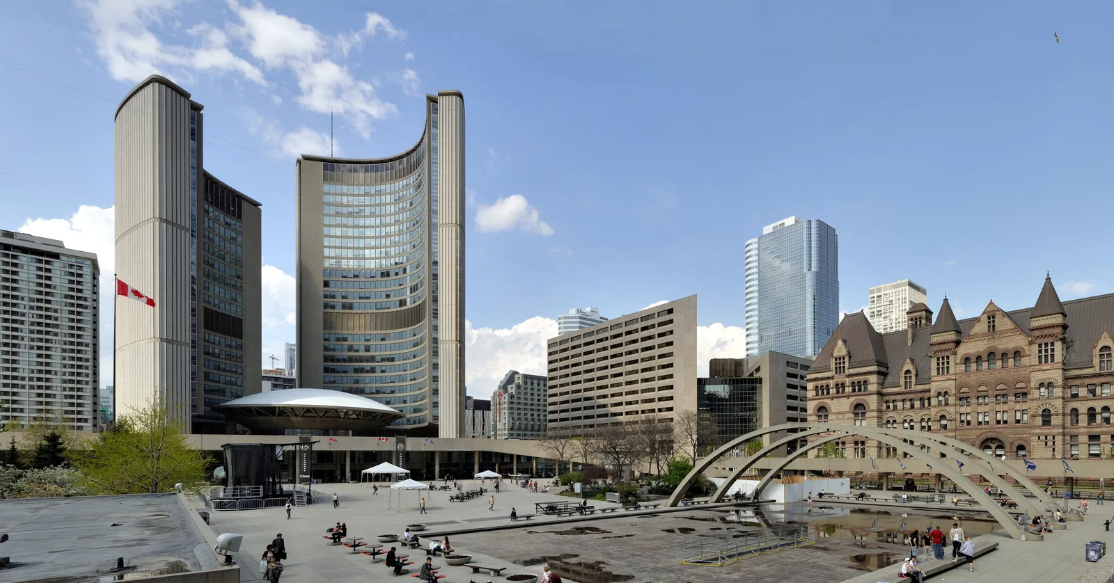 Toronto City Hall seen across Nathan Phillips Square, two curved concrete towers of different heights standing around a low saucer-shaped council chamber, with the square's concrete arches and reflecting pool on the right