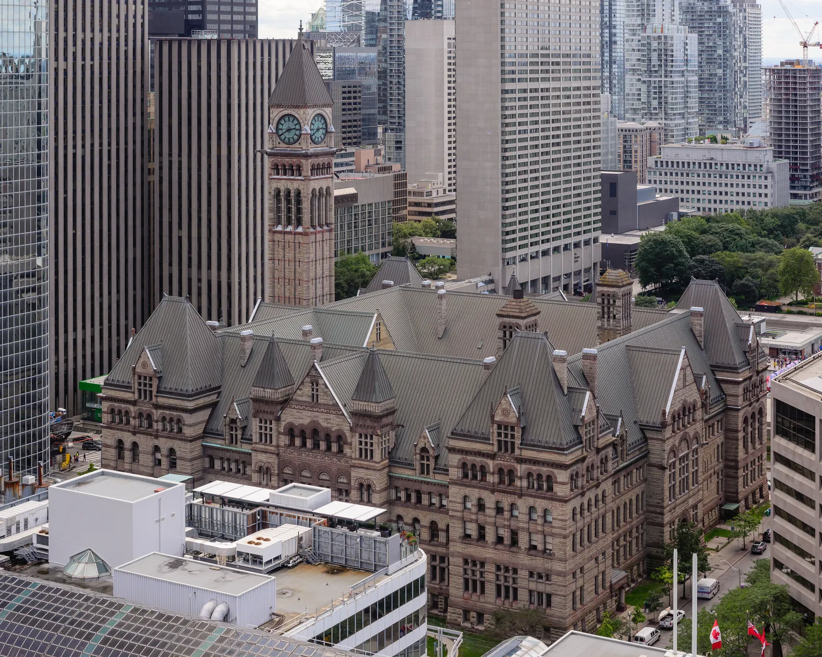 Toronto's Old City Hall seen from above, a long sandstone building with steep slate roofs, dormers and turrets, its clock tower rising off-centre against the glass towers of the financial district behind