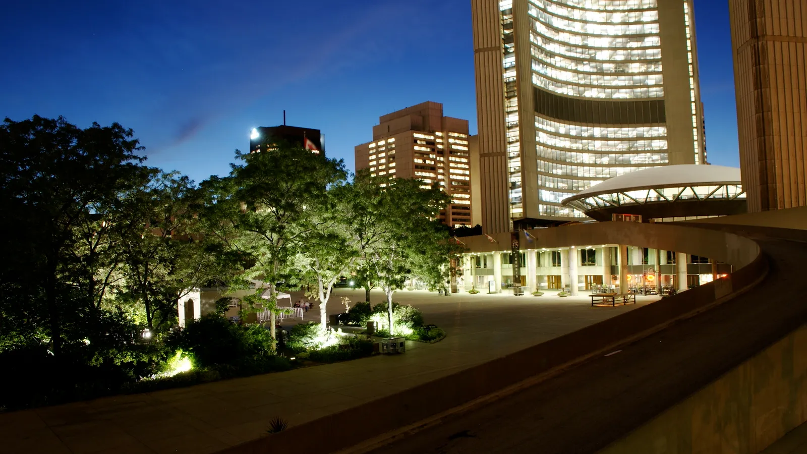 The Peace Garden beside Toronto City Hall after dark, its trees and paths lit from below, with the curve of the city hall tower and the roof of the council chamber behind them