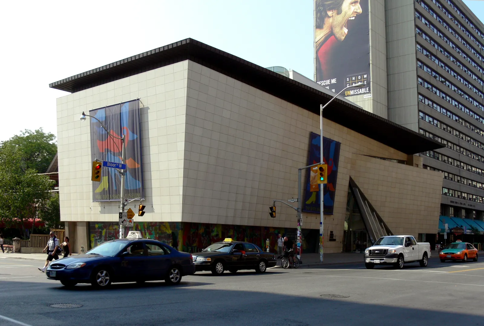 The Bata Shoe Museum on Bloor Street West in Toronto, a pale limestone block with an overhanging angled roof like the lid of a shoe box, banners hung on its corner and traffic passing in front