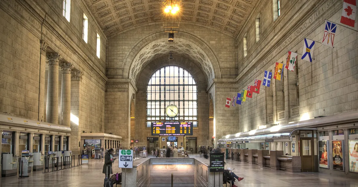 The Great Hall inside Toronto's Union Station, a long stone chamber under a high tiled vault with tall arched windows at the far end, provincial flags strung along the right-hand wall and departure boards below them