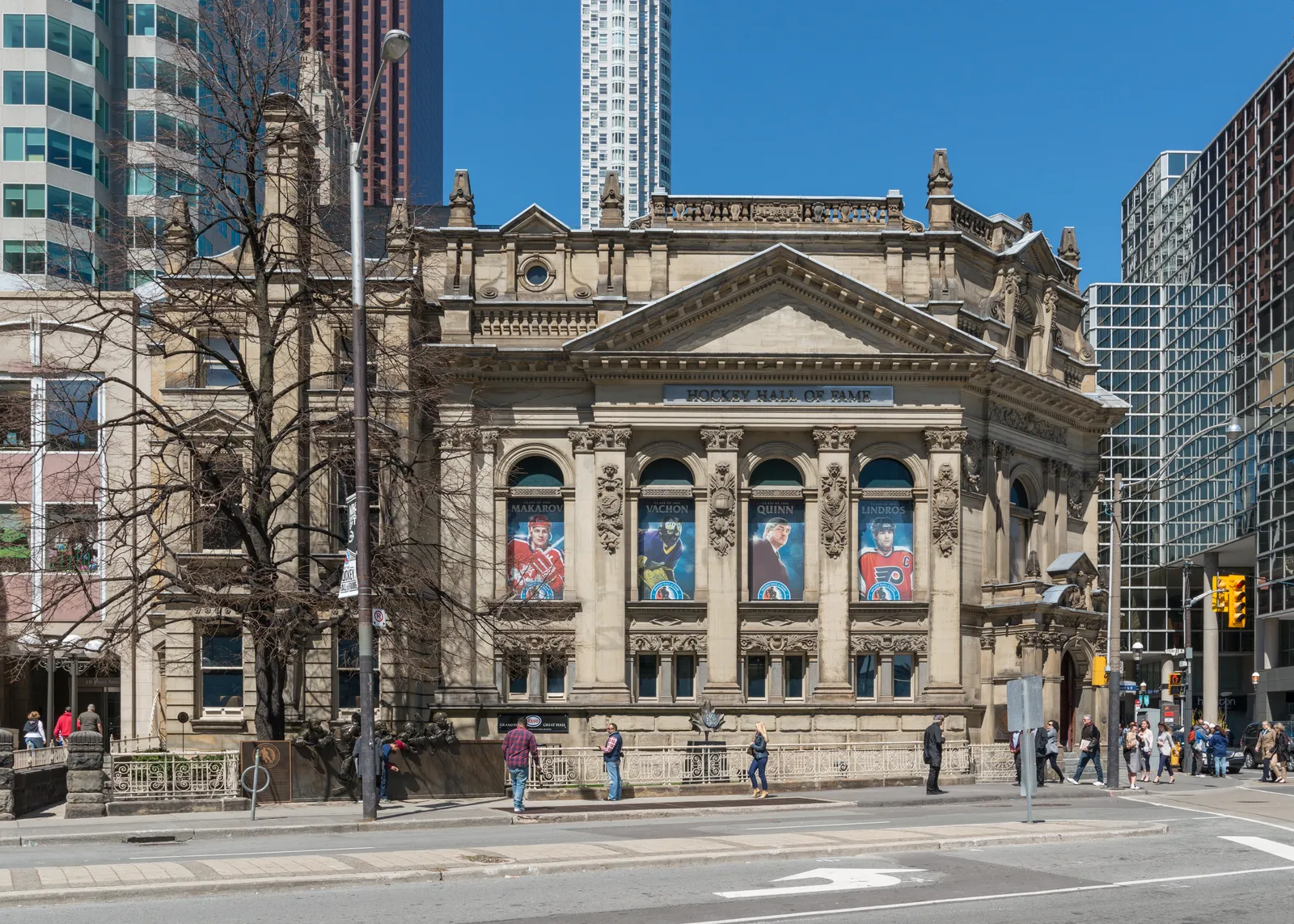The Hockey Hall of Fame in Toronto, an ornate carved stone bank building of 1885 with a pediment and columned windows carrying hockey banners, glass office towers rising directly behind it