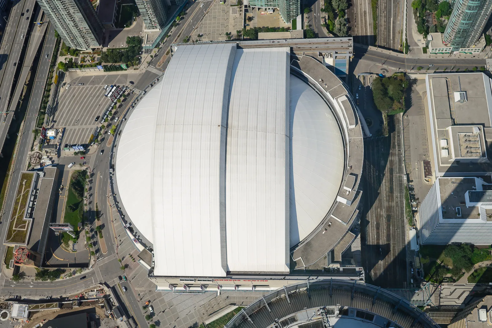 The white domed roof of Rogers Centre in Toronto seen from directly above, the seams between its sliding panels visible across the top, with rail tracks and city streets around it