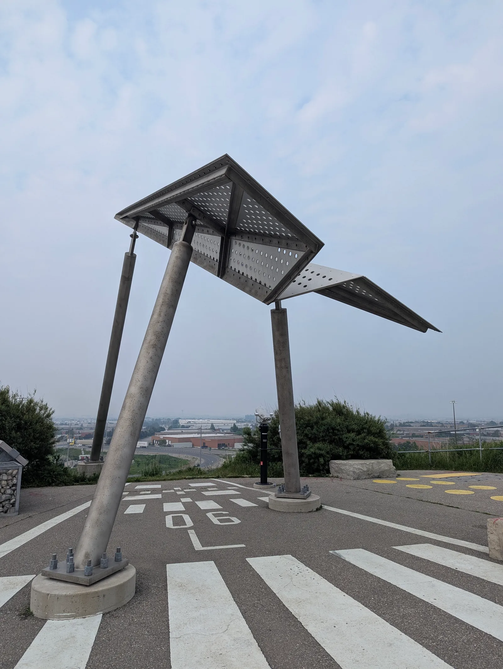 The airplane viewing platform at Danville Park in Mississauga, a perforated steel wing-shaped canopy on angled concrete legs standing over paving marked with runway-style white stripes and numbers