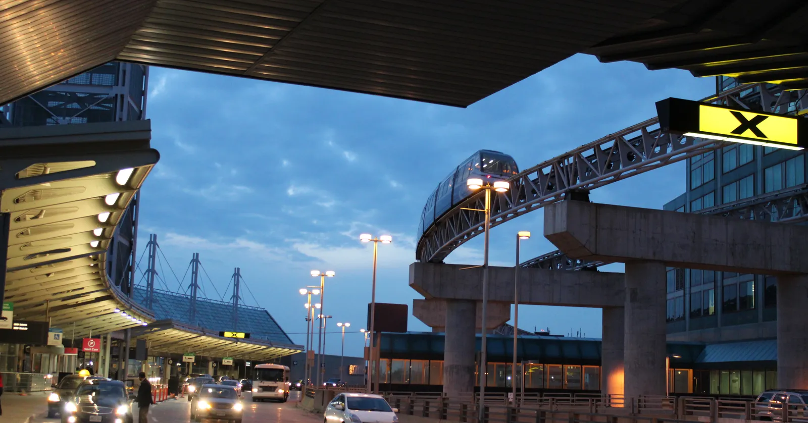 The departures curb at Toronto Pearson Terminal 1 at dusk, with the terminal's long curved roof on the left and a Link train on its elevated concrete guideway crossing the road on the right