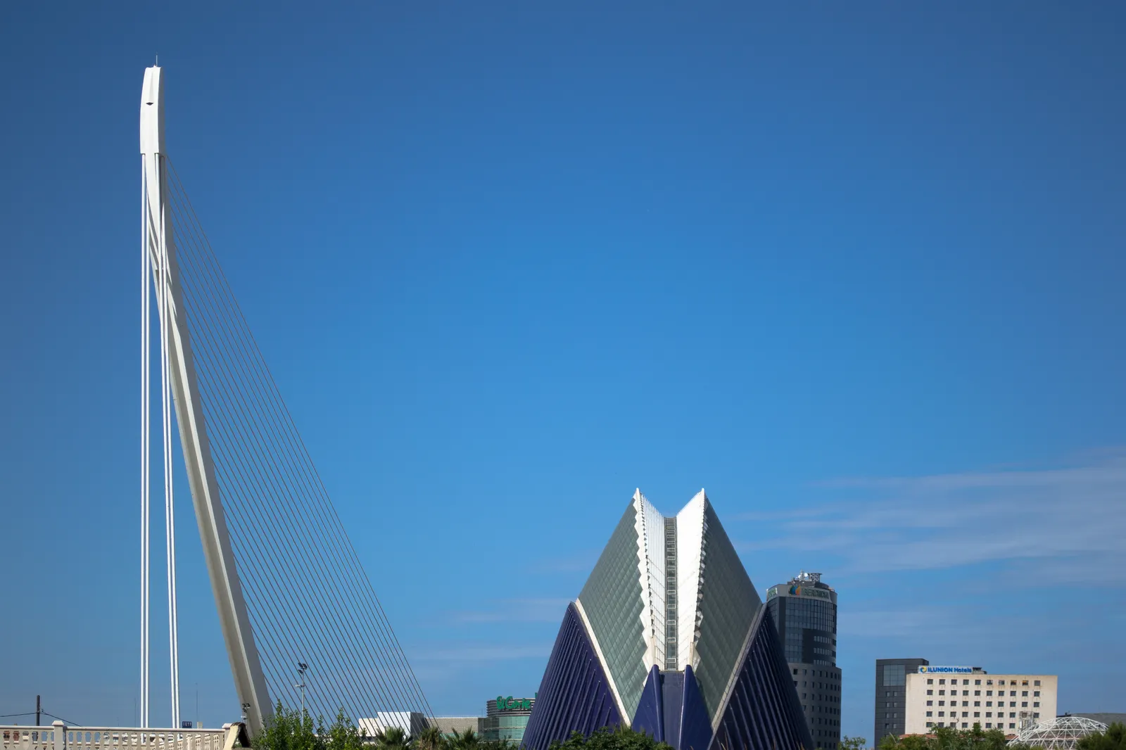 The white single pylon of the Assut de l'Or bridge in Valencia rising against a clear sky, with the folded roof of the Àgora beside it