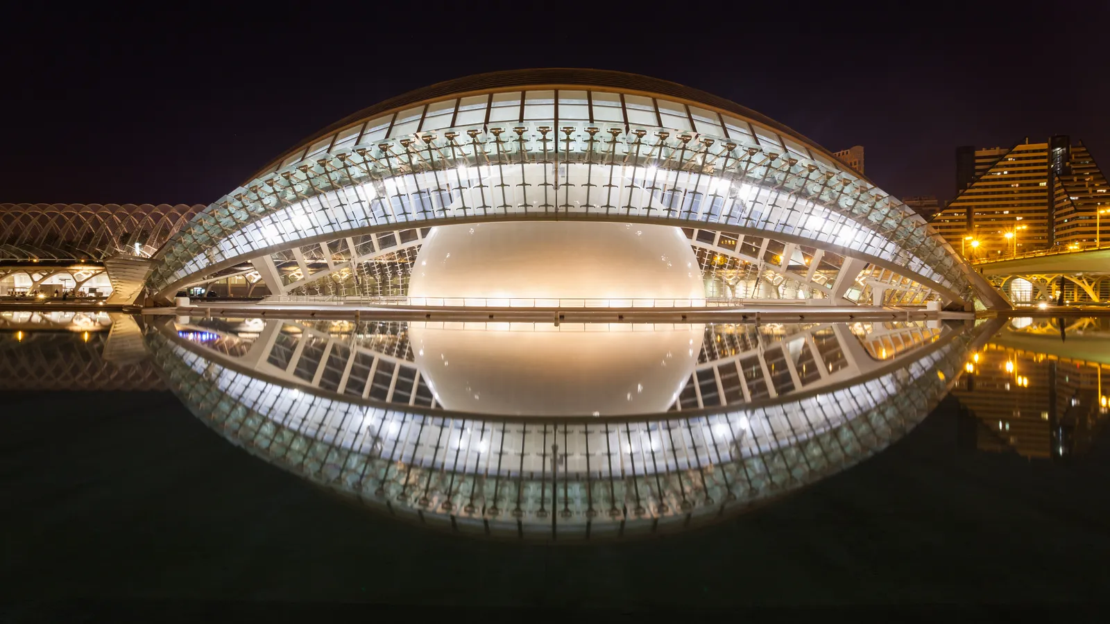 The Hemisfèric in Valencia lit at night, its glazed dome and curved roof mirrored in the pool below so the building and its reflection together form an eye